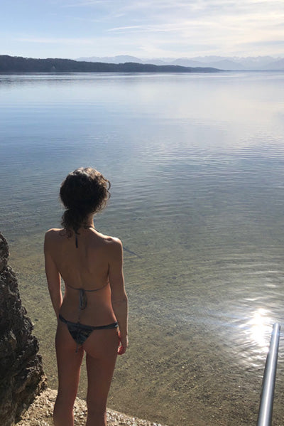 Yoga Lehrerin Ranja Weis im Bikini steht am Ufer eines Sees und blickt auf das Wasser.