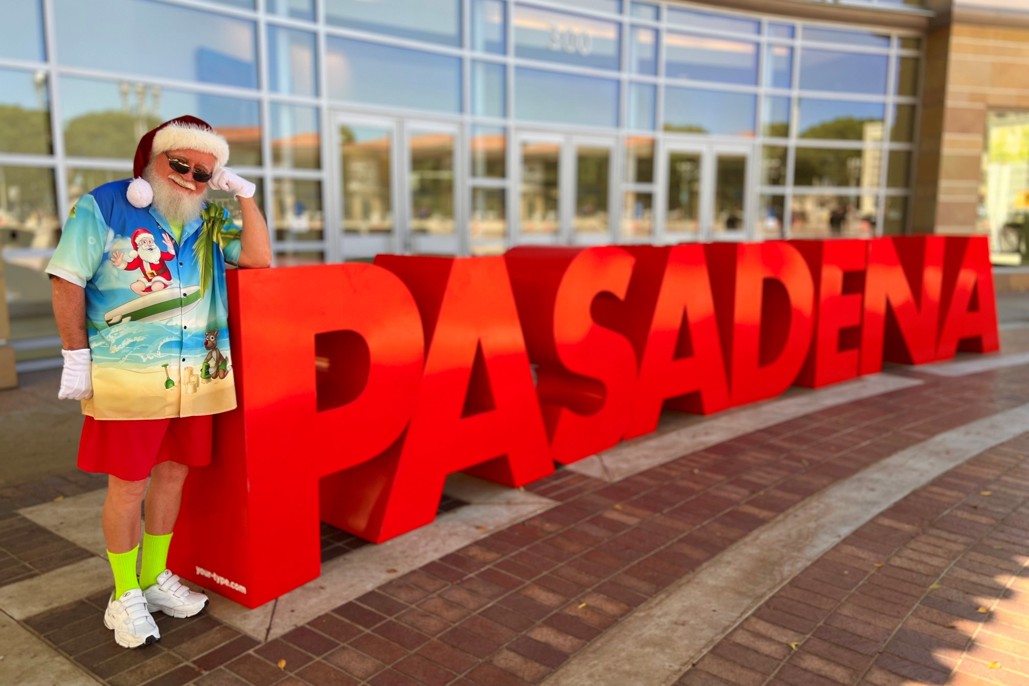 Santa in a beach-ready summer outfit standing in front of a sign that says Pasadena at Christmas Con CA 2022 in California.