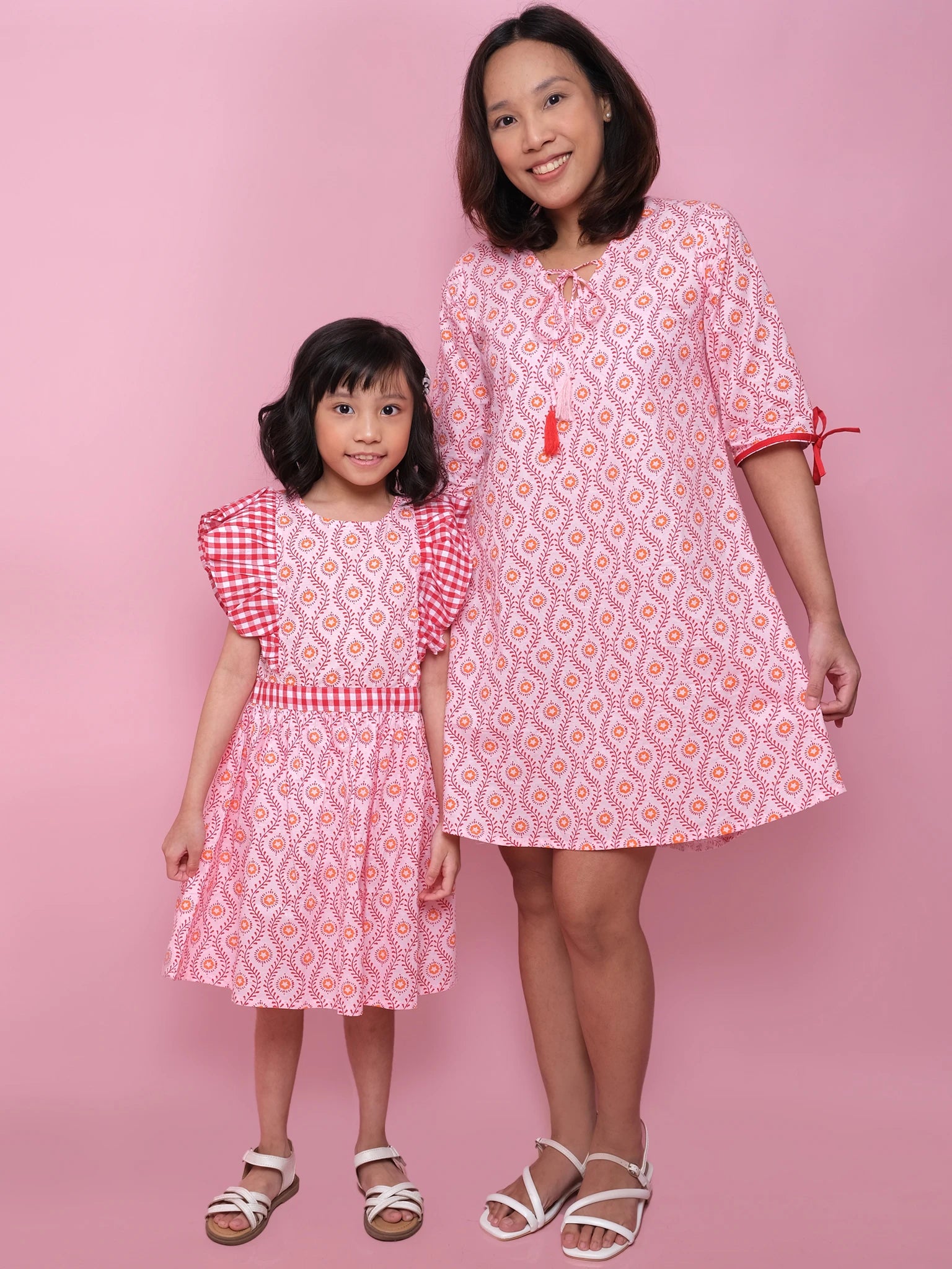 An adult and child stand side by side against a pink background, both in matching Woodblock Flower Jayne Dresses by Woodblock Flower, featuring woodblock print patterns and white sandals.