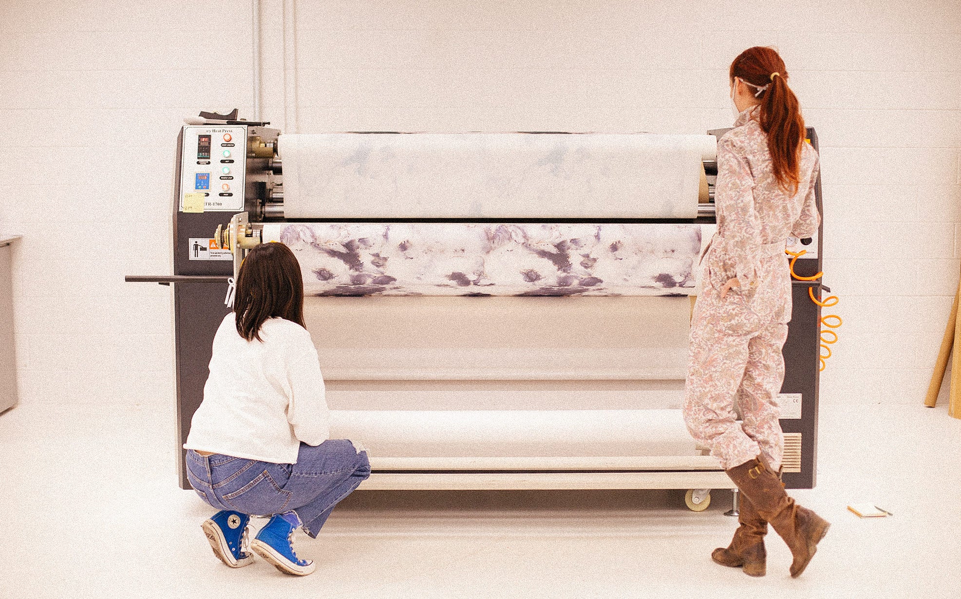 Two women are in front of a large fabric printed. The woman on the left is wearing blue sneakers and kneeling while the woman on the right wears tall boots and stands with her foot kicked back and a hand in her pocket.