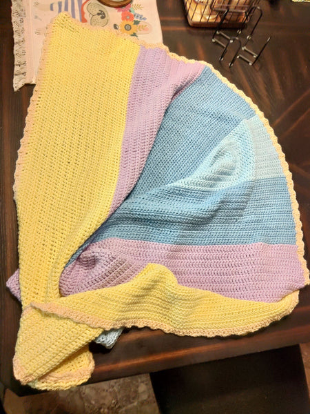 A baby blanket is arranged on a dark table.  The blanket is yellow green, purple, blue, and light blue with a yellow edging.