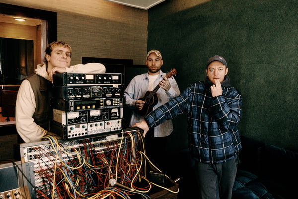 Three members of DMA's stood next to soundsystem equipment