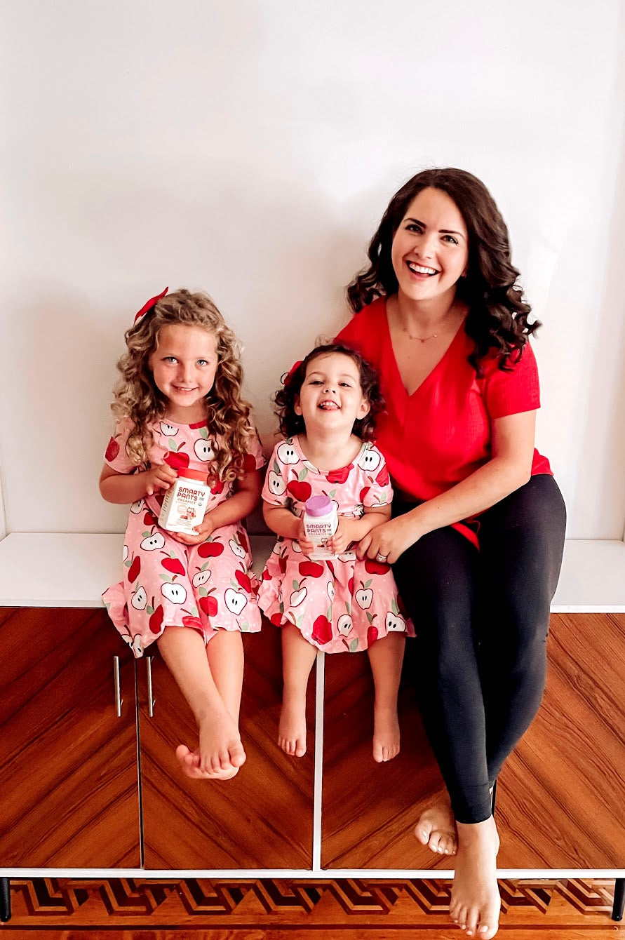 Woman sits on bench at home with her two little girls in matching apple print dresses. The girls are holding SmartyPants vitamins.