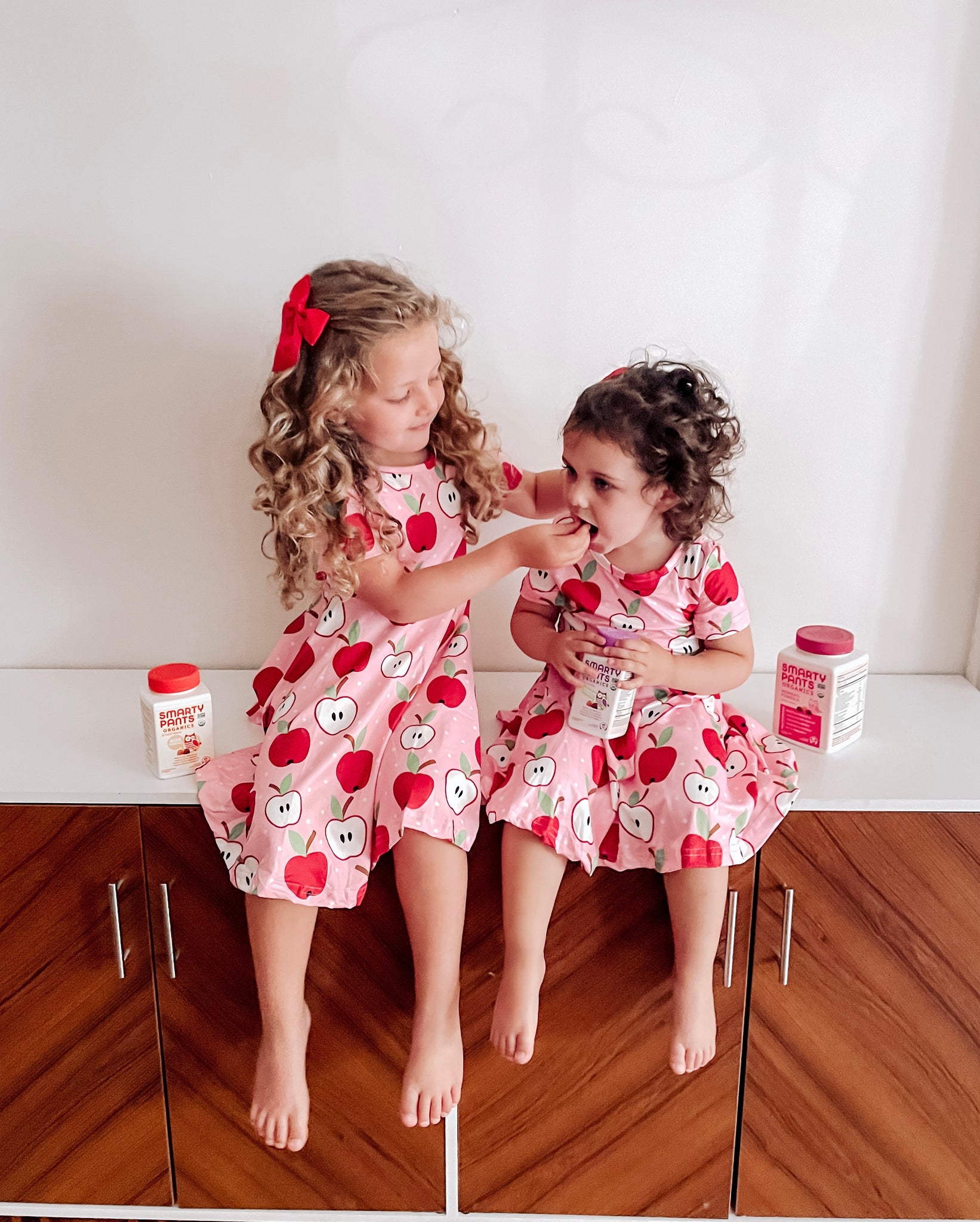 Two little girls sit inside on a bench, in matching apple print dresses. One little girl is feeding the other a SmartyPants Vitamins gummy.