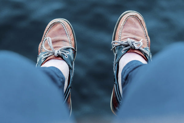 A pair of legs hanging from a boat with the sea in the background. In focus, a pair of boat shoes wore by the model.