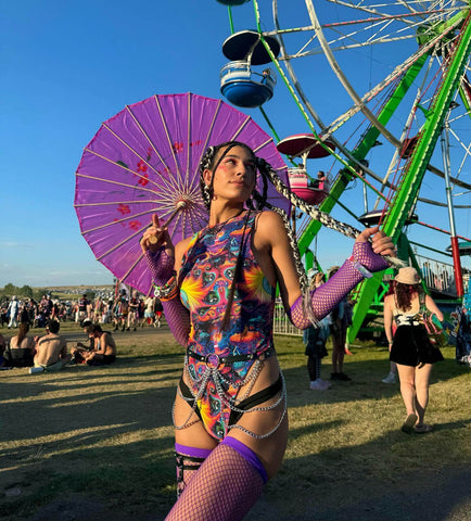 A woman showcases vibrant rave apparel, featuring a colorful bodysuit, purple fishnet gloves and stockings, accessorized with a purple parasol and braided hair, embodying the energetic festival spirit