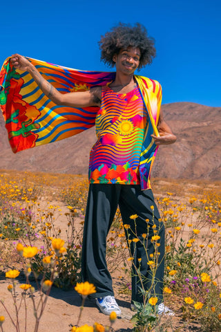 A person wearing a colorful, psychedelic rave outfit, standing in a field of yellow flowers with mountains in the background