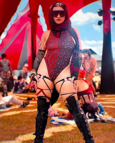 A festival-goer in a red and black geometric-patterned bodysuit, fishnet sleeves, strappy leg harness, and black boots poses confidently.