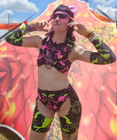 A festival-goer in a vibrant women's rave outfit poses confidently. She wears a neon pink and green patterned set with matching accessories and sunglasses, set against a colorful festival backdrop