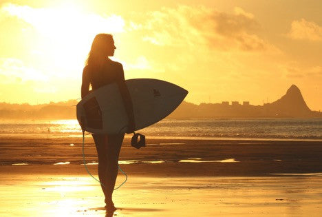 An athletic young woman walks toward the setting sun while holding a surfboard. This is the lead image in H2O Audio's Mother's Day gift guide.  