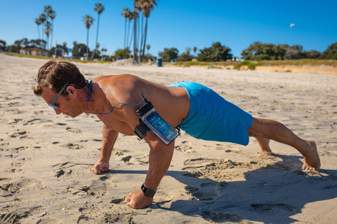 An athletic man does pushups on the beach while wearing the Amphibx waterproof case on his upper arm. He's also using the H2O Audio Surge waterproof headphones to listen to music while he works out on the beach. 