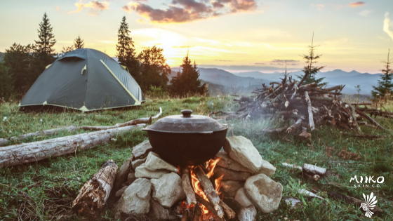 image of a campsite up on a mountain. There is a pot cooking over a fire in the foreground, and a tent set up in the background. The whole campsite is overlooking a beautiful mountain valley and the sun is setting in the background