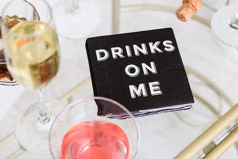 Black paper beverage napkins with white, bold text that reads, "Drinks On Me"