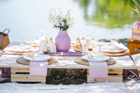 picnic set up with white plates and bamboo placemats.  Purple vase in the center has white flowers .  Set up overlooking the pond