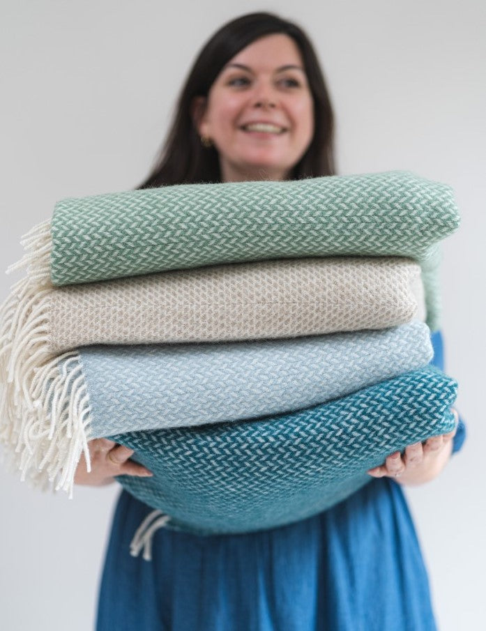Smiling woman holding a stack of blue and green wool blankets and throws from The British Blanket Company