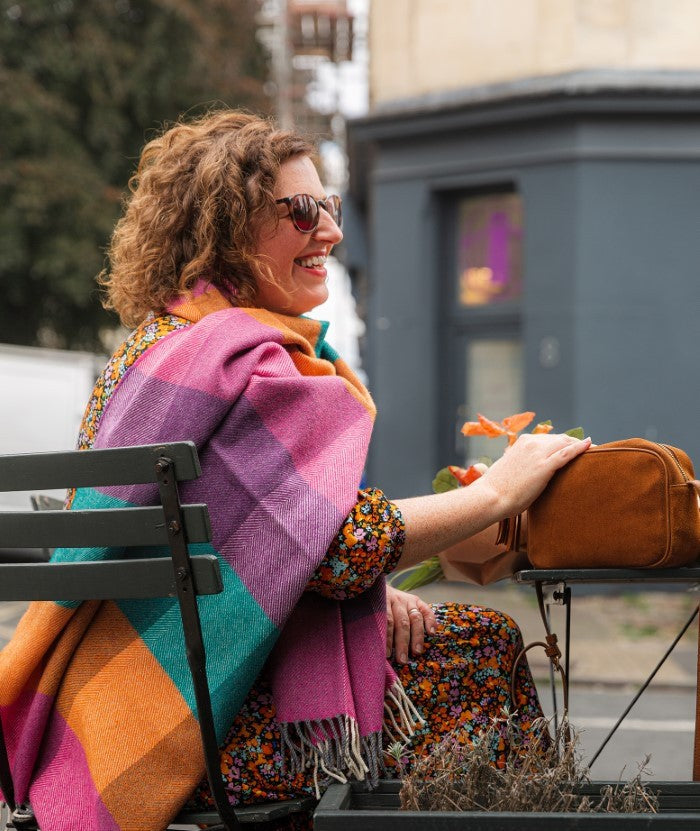 Smiling woman sitting outside a coffee shop wearing a multicoloured scarf from The British Blanket Company