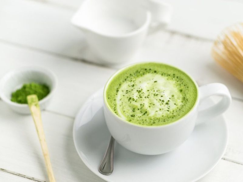 A matcha latte in a white cup with milk foam with matcha powder and bamboo whisk in the background