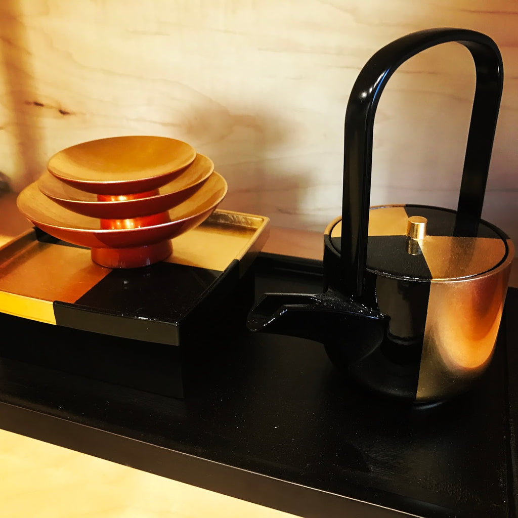 A photo of a black and gold lacquered Japanese sake set which consists of a serving pot, two dish-like cups, and a tray