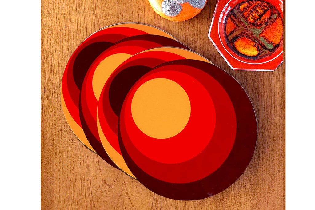 A set of four circular placemats featuring 70s style graphics lie in an overlapping line on a teak table