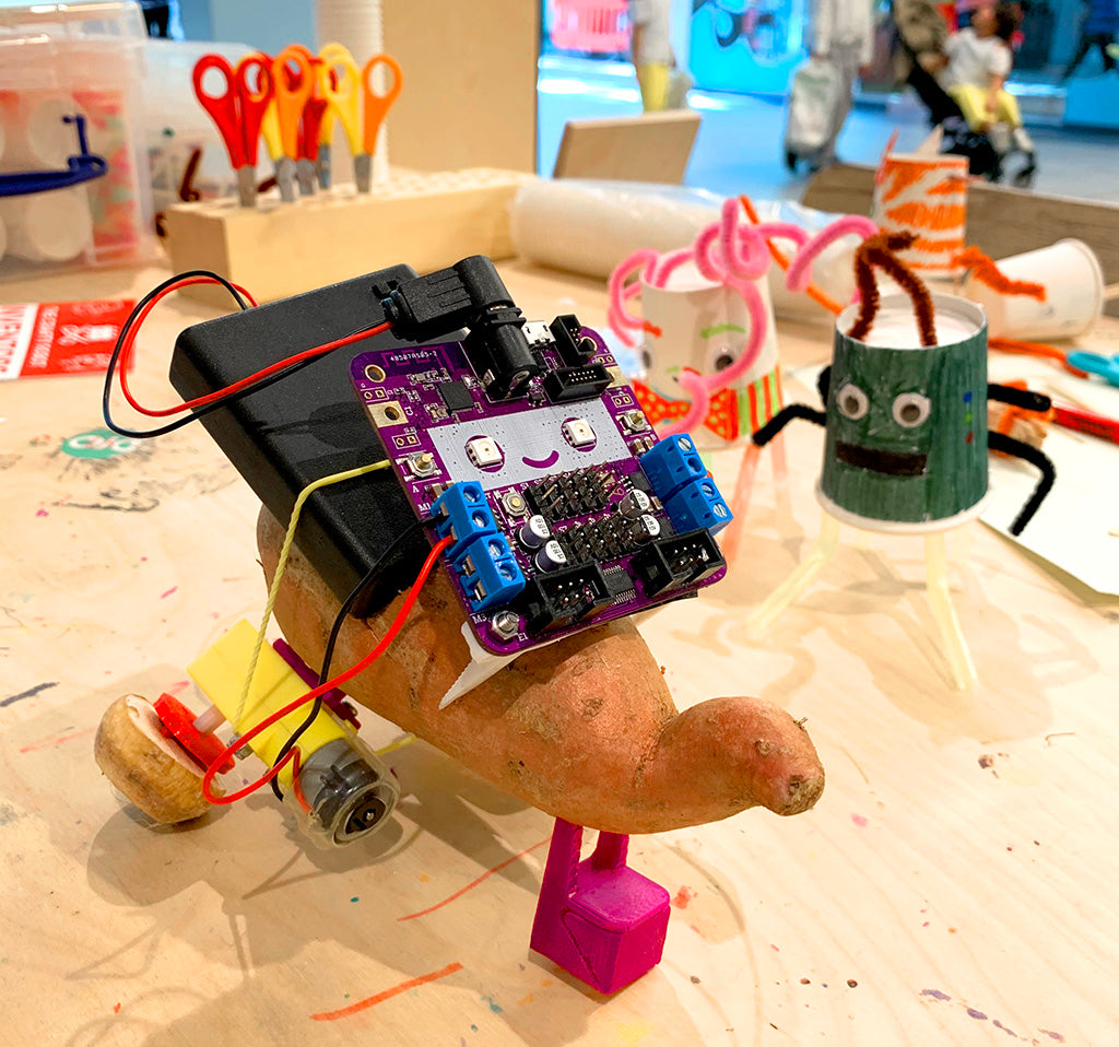 Photo of a robot with a body made from a sweet potato and wheels made from parsnip slices. On the front is a square purple circuit board with smiley face.