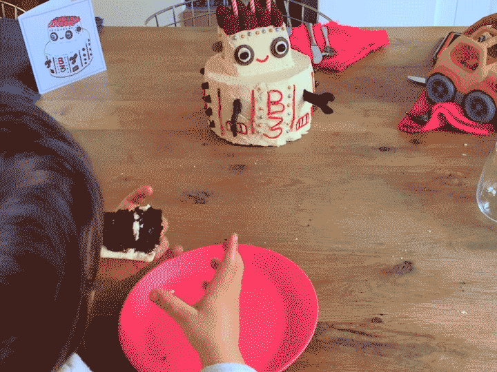 Looping video of a robot cake driving around on a table. In the foreground a toddler eats a piece of the cake