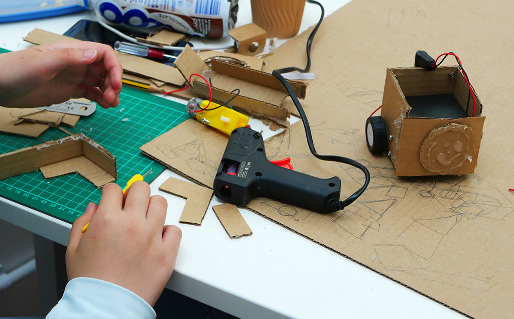 Photo of a partially completed cardboard robot with some intricate cardboard pieces being assembled with a hot glue gun in the foreground. A motor and some tools lie in the background.