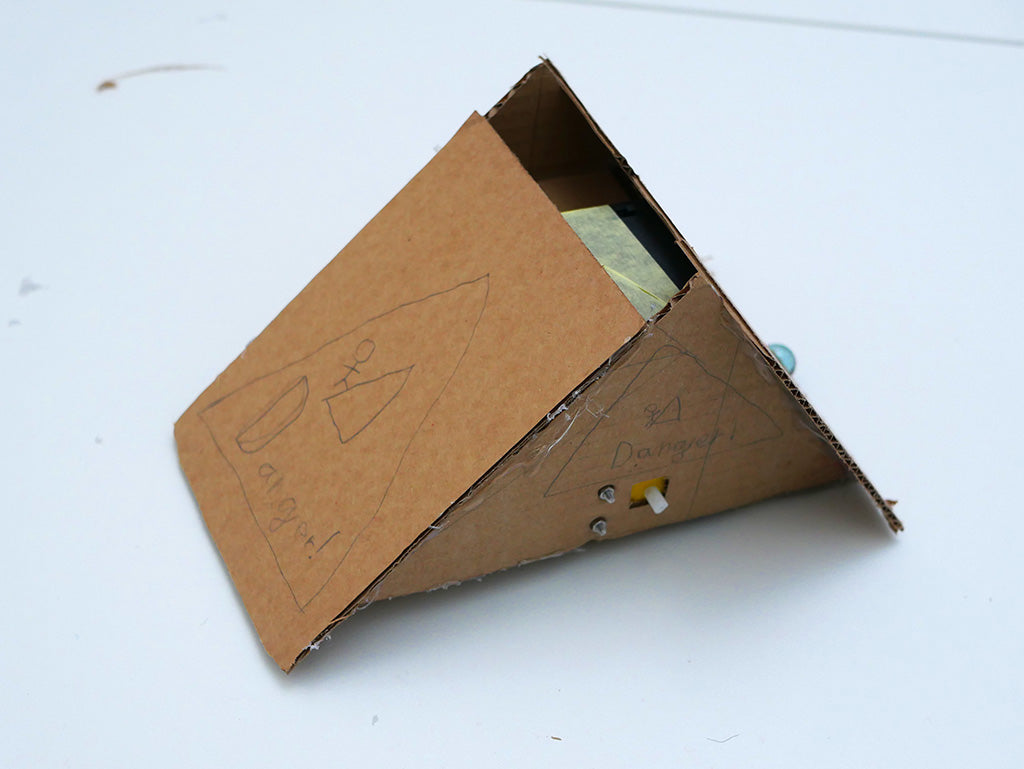 Cardboard robot in the form of a wedge, decorated on the front and side with pencil drawings of a stick man being attacked by a triangle and the word 'Danger!' inside a triangle. 