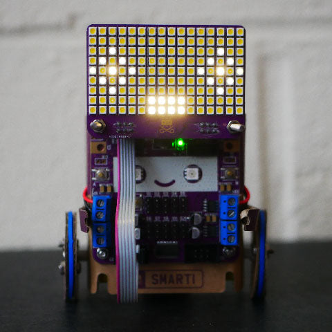 Smartibot with an LED matrix attached to the top displaying a face with dull eye outlines, bright pupils and a bright, rectangular mouth