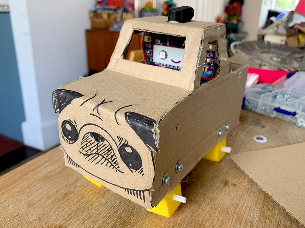 Photo of a cardboard monster truck with a purple smiling circuit board in the cab, four yellow gear motors and no wheels