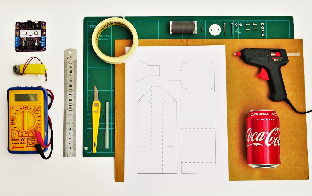 Photo showing tools and materials including circuit board, multimeter, motor, screws and bolts, coke can, pulley, Sellotape, glue gun, ruler, string, cardboard sheet, paper template