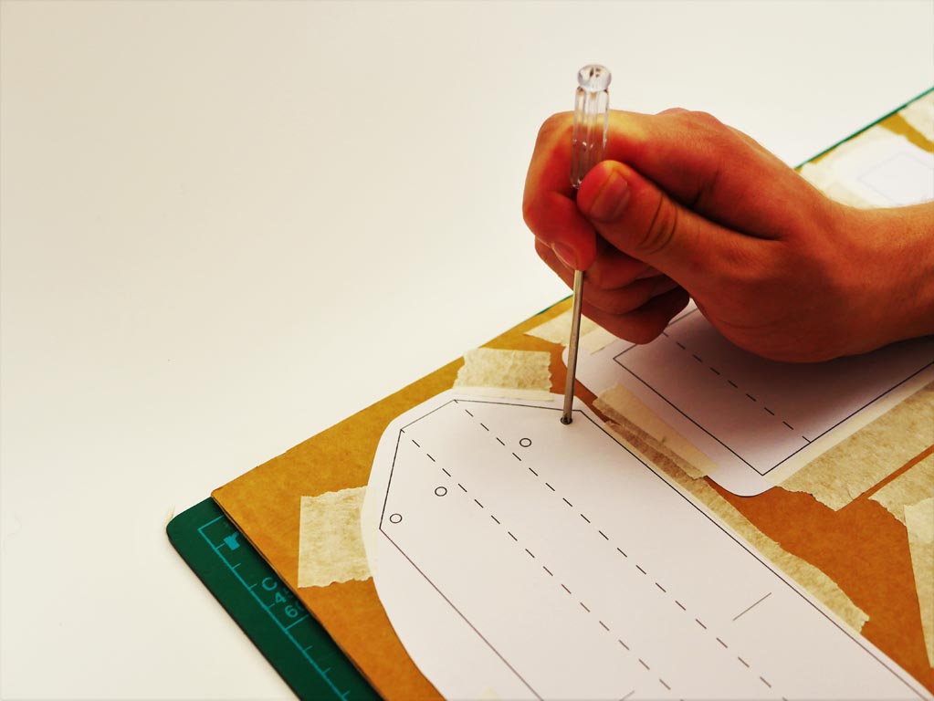 Image shows screwdriver being used to poke holes through the cardboard at the points where the paper templates indicate.