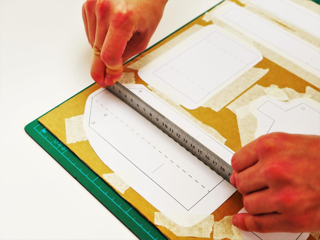 Image shows a ruler being pressed down along the dotted lines on the paper template in order to create edges for which the cardboard to fold around later on.