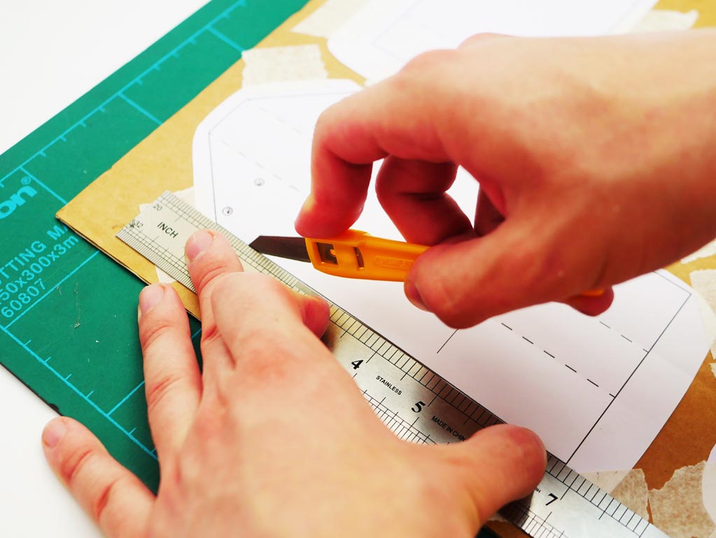 Image shows a craft knife with a ruler being used to cut out the cardboard shapes along the template lines.