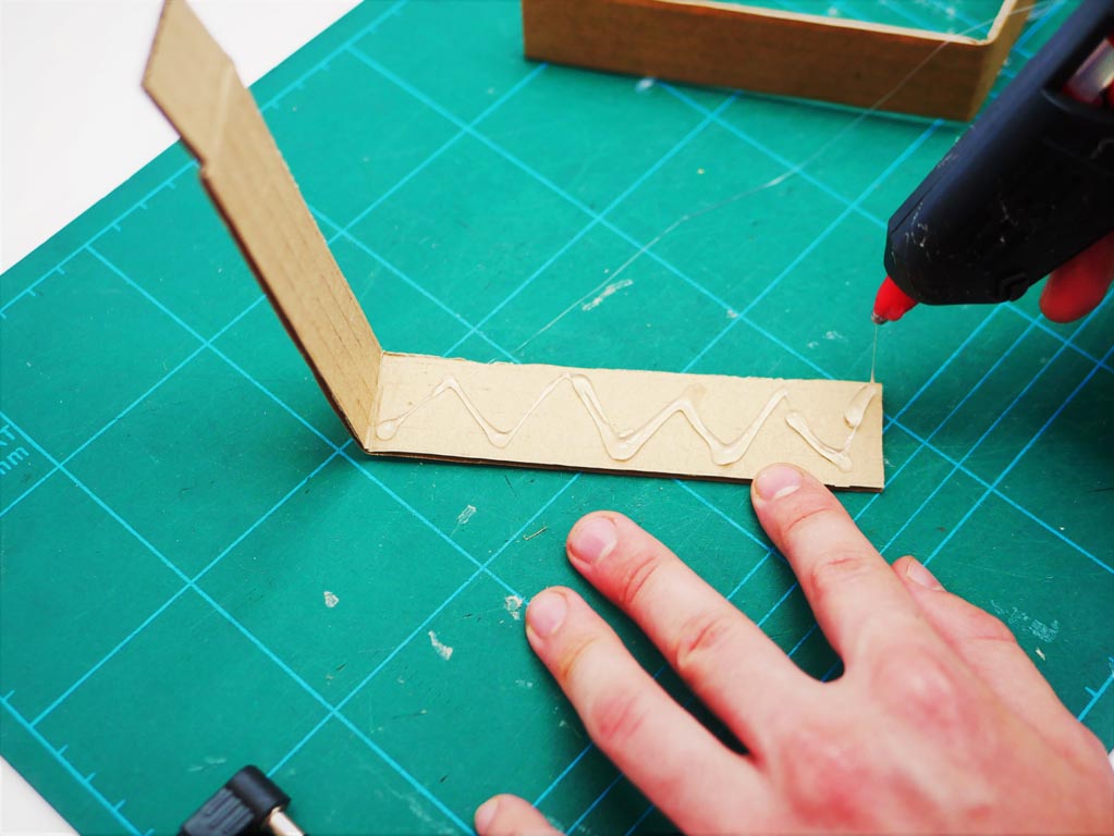 Image shows glue gun being used to spray a zig zag pattern onto cardboard  back frame of the crane.