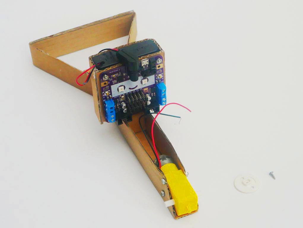Image shows circuit board now screwed onto box on crane with battery box fitting just inside of the same cardboard box, with pulley and screw adjacent.