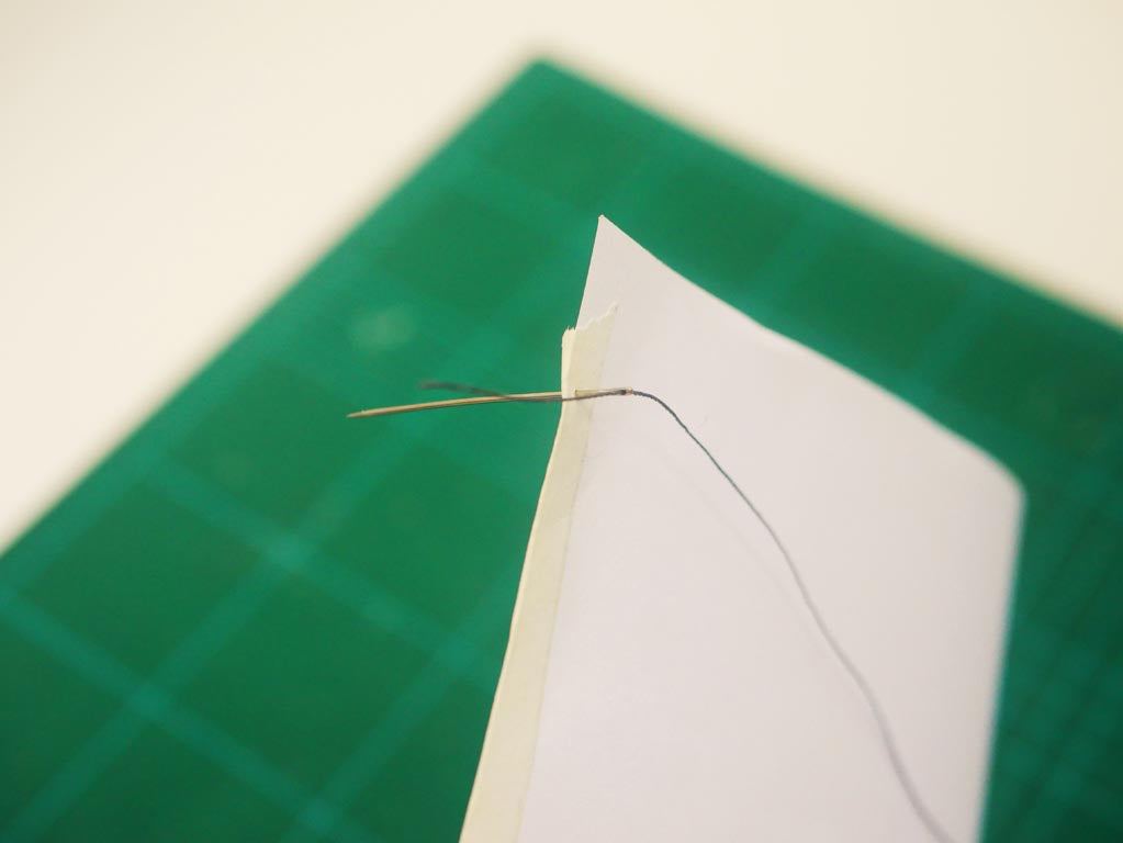 Image shows needle with string on it being put through top of a4 paper where it is folded.