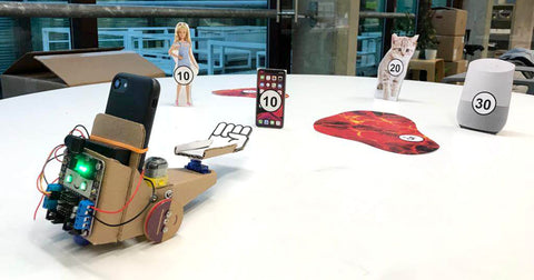 Photo of a small cardboard robot carrying a smartphone and with a motorised cardboard fist on the front on a table on which there are some cardboard cut out items like a Barbie, a kitten and an iPhone and some patches of lava. All the items and lava have circles with numbers in them