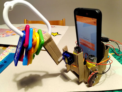 Photo of a cardboard robot carrying a smartphone. On the front of a robot a brightly coloured baby toy is attached to a cardboard arm which is attached to a small servo motor.