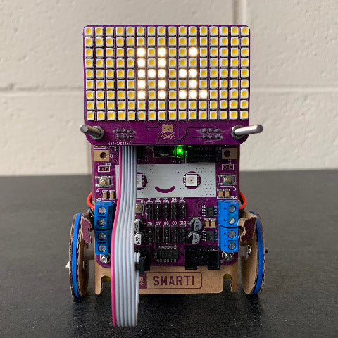 A small cardboard robot (Smartibot) with an LED matrix on the top displaying the text 'Hi'