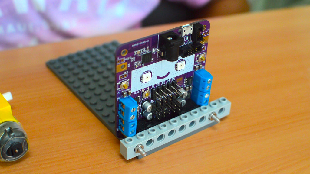 Image of a purple circuit board with a smiling face screwed onto a light grey LEGO Technic brick
