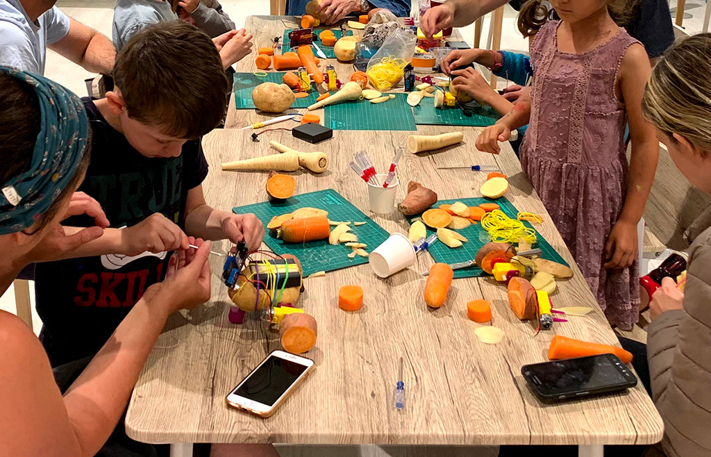 A table covered in pieces of vegetables, electronics and smartphones with children and adults intently working