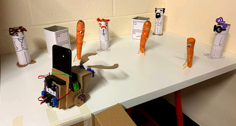 Photo showing a small cardboard robot carrying a smartphone and with a cardboard grabber claw on the front with some figures made from carrots and sticks and some more made from cardboard and pipe cleaners arranged in front of it on a table 