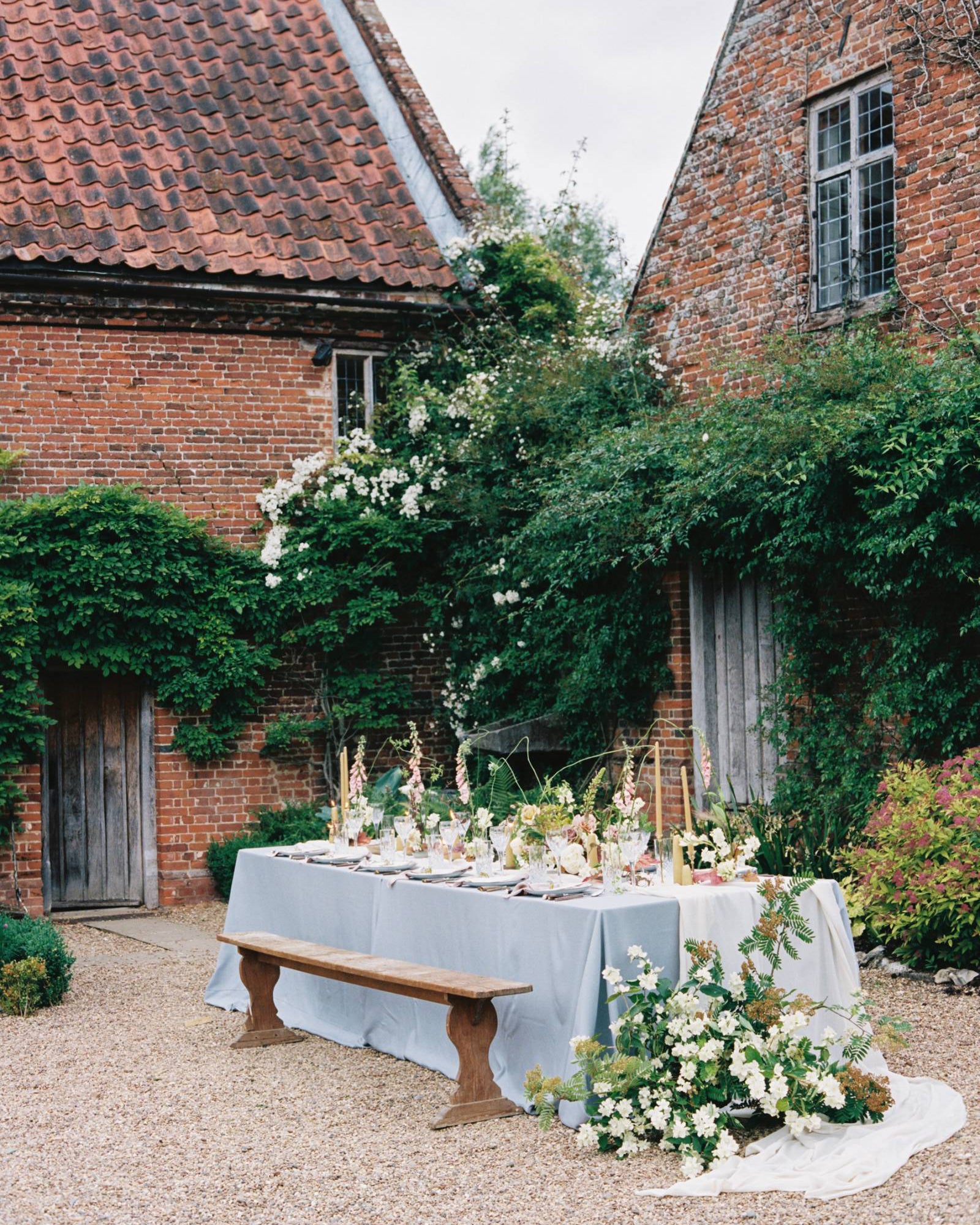 Outdoor table setting with a white tablecloth covering the table, large floral arrangements and beeswax candles. Shown in a courtyard surrounded on both sides by a period home.