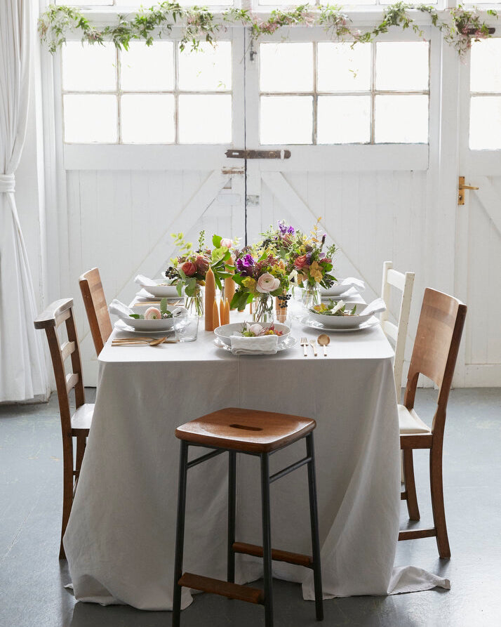 Table setting with an oversized white tablecloth, wooden chairs, floral arrangement and beeswax candles.