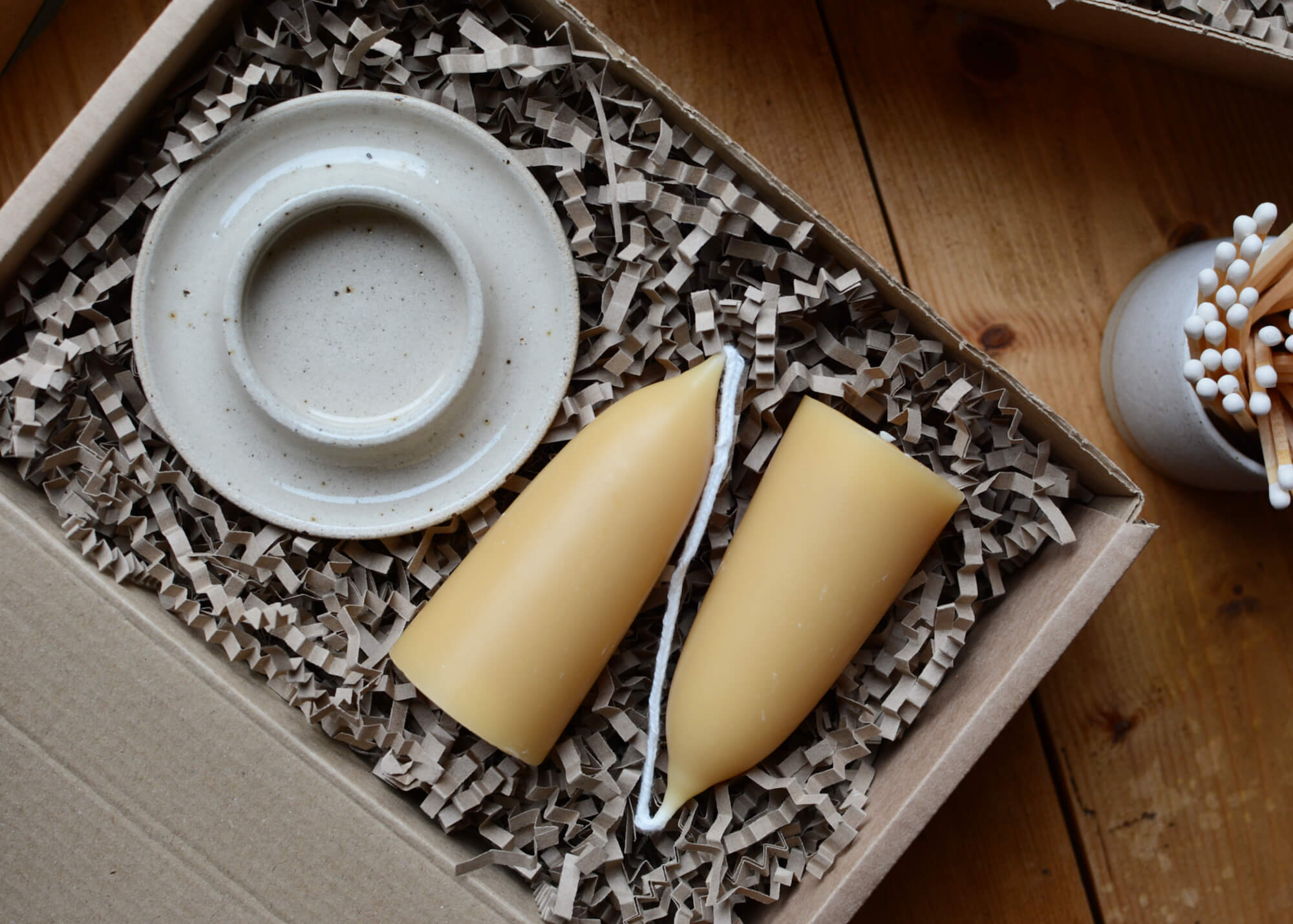A pair of beeswax pillar candles joined at the wick alongside a ceramic candle holder, presented in a kraft gift box