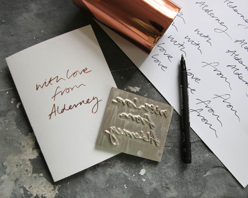 a hand foiled card with the message With Love From Alderney on it surrounded by rose gold foil and the paper the phrase was tested on before the metal die was used.