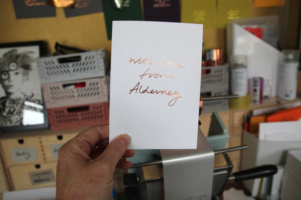 hand holding a white card with the phrase with love from alderney handwritten and hand foiled on the front
