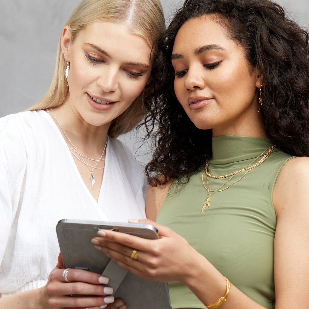 Two women wearing the FIYAH plume jewellery collection in silver and yellow gold plated while looking at a picture of a loved one in a photo frame