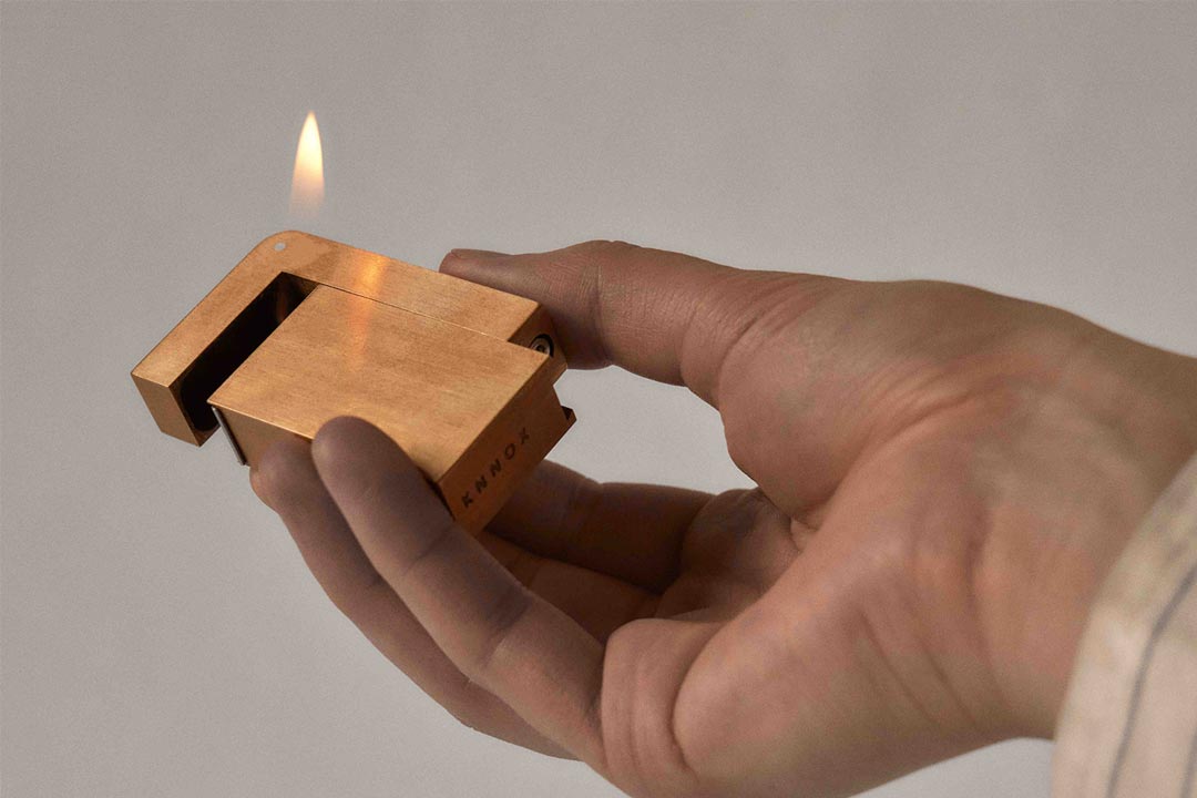 A square brass cigarette lighter with flame being held open by a hand