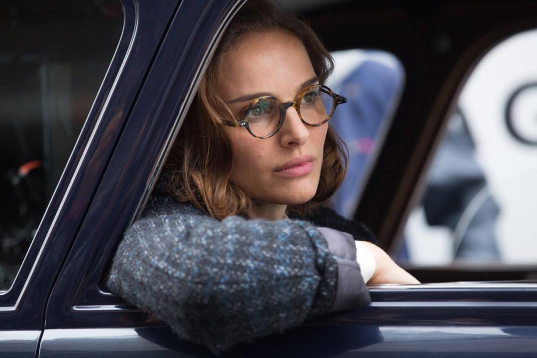 Actress Natalie Portman leaning out of blue car window wearing thick jumper and round tortoiseshell spectacles